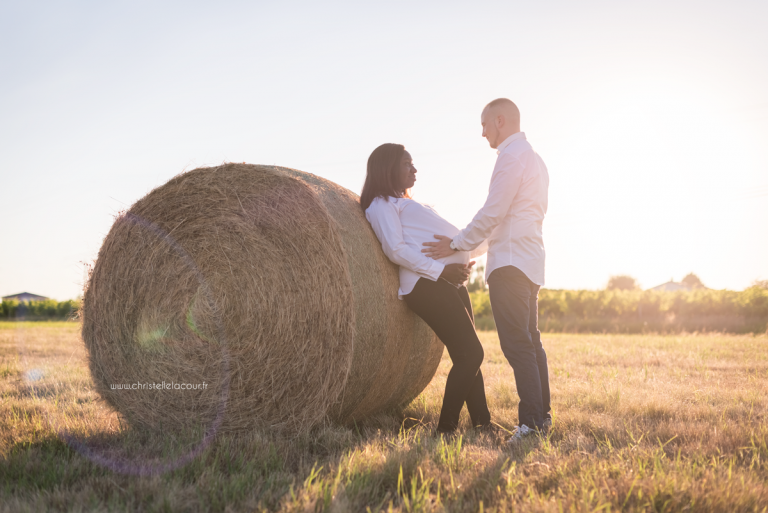 Photographe de grossesse à Toulouse, couple de futurs parents adossés à une botte de paille au coucher de soleil